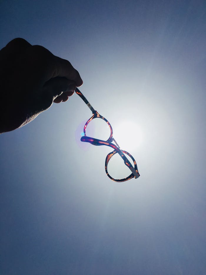 A hand holds colorful glasses against the bright sun creating a striking silhouette.